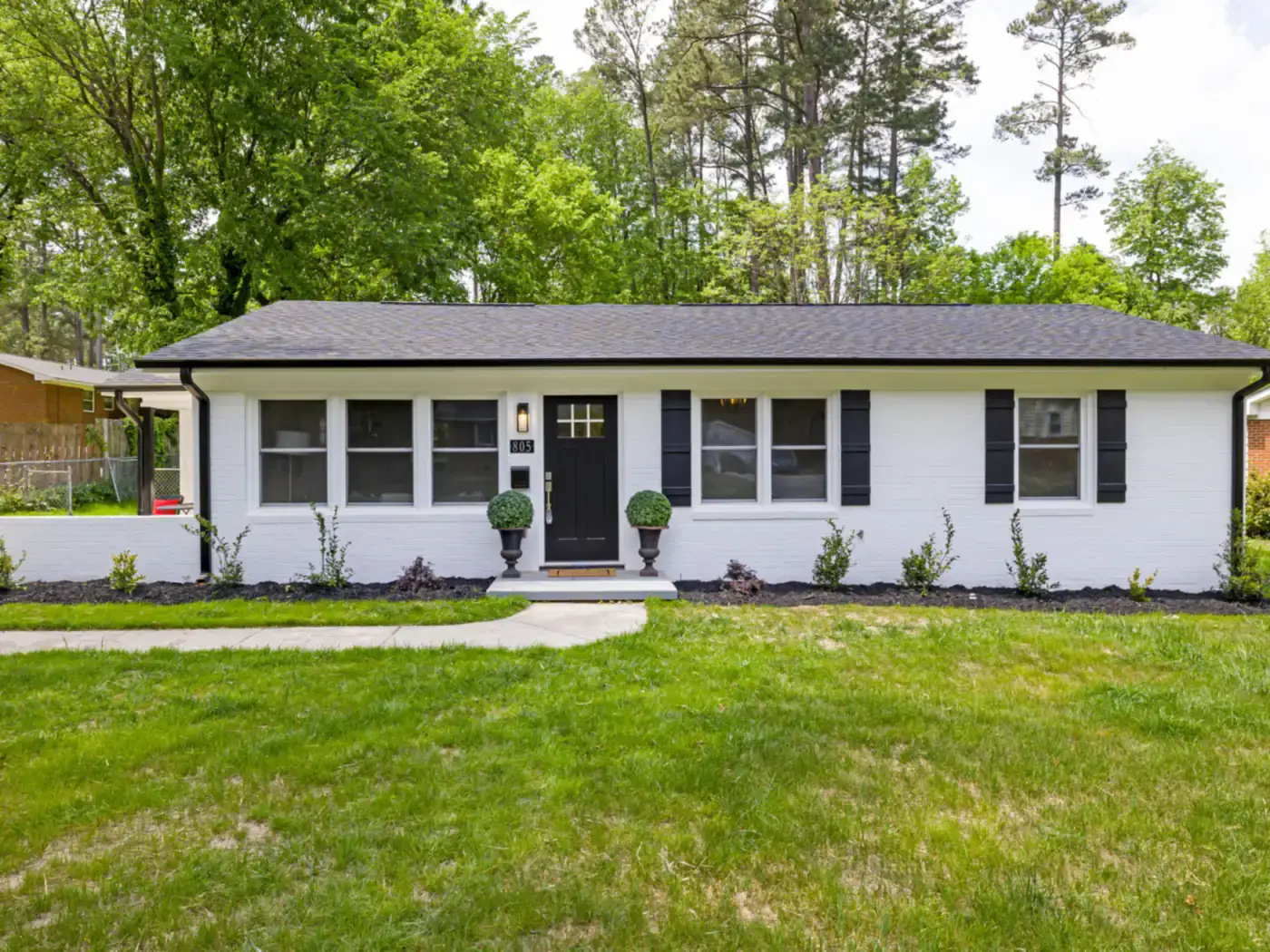 White single-story ranch home with black shutters, a green lawn and tall trees behind it