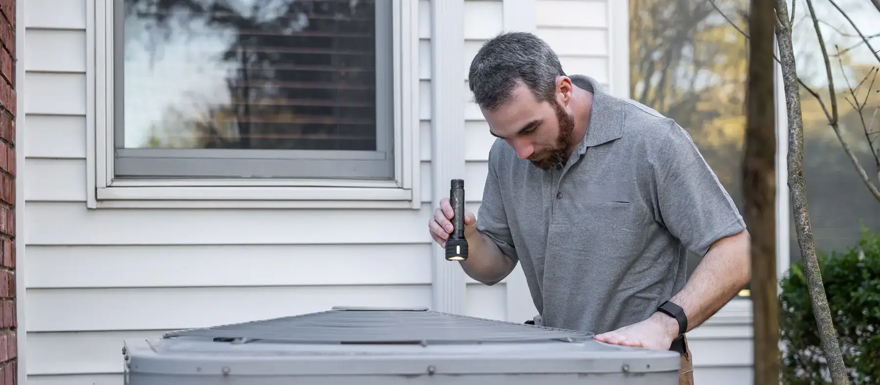 Technician with a flashlight inspecting an outdoor air conditioning unit beside a house