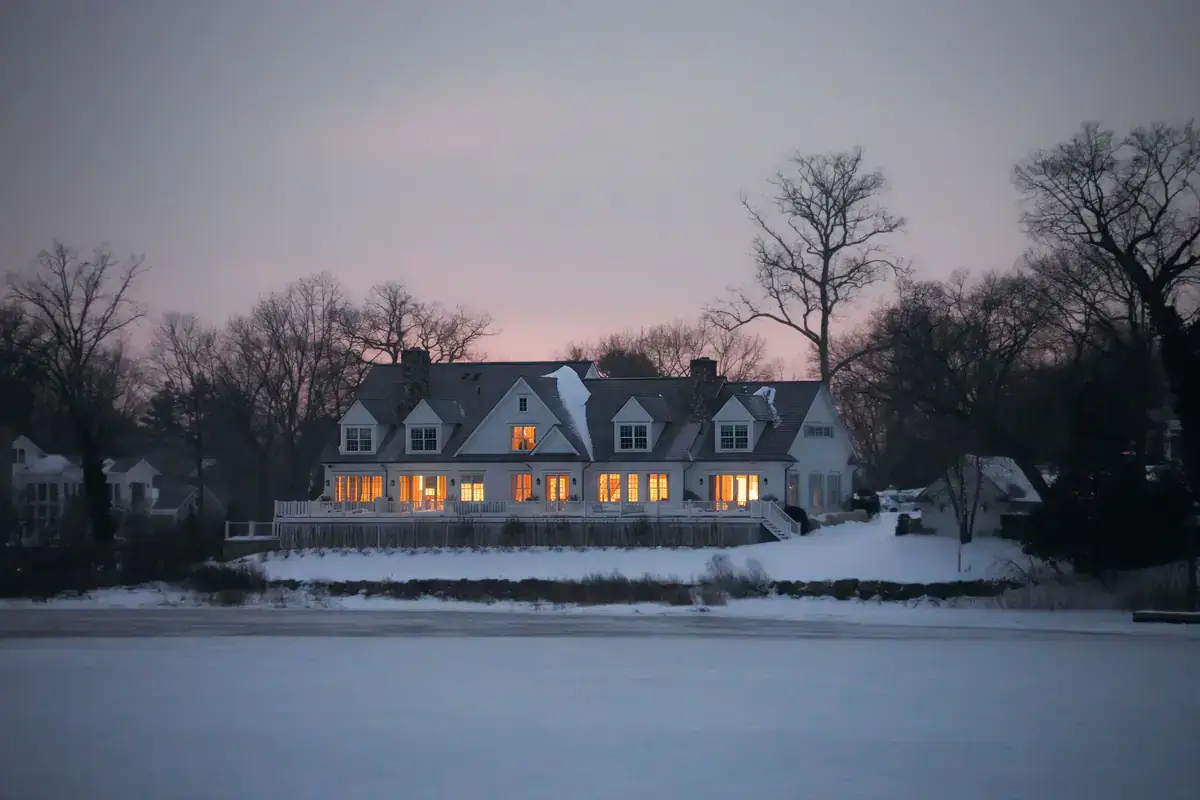 Large house with lit windows across a snowy field at dusk