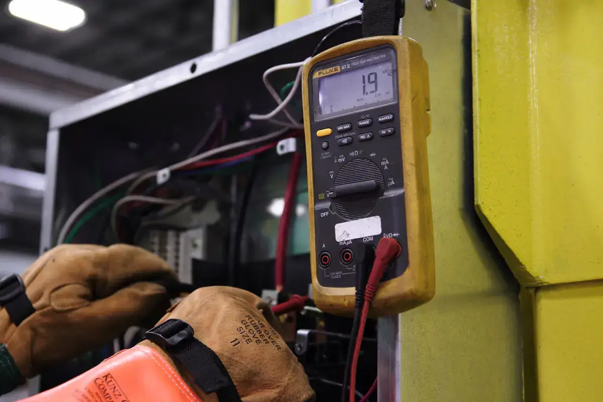 Gloved hands testing wiring inside an equipment control panel with a digital multimeter