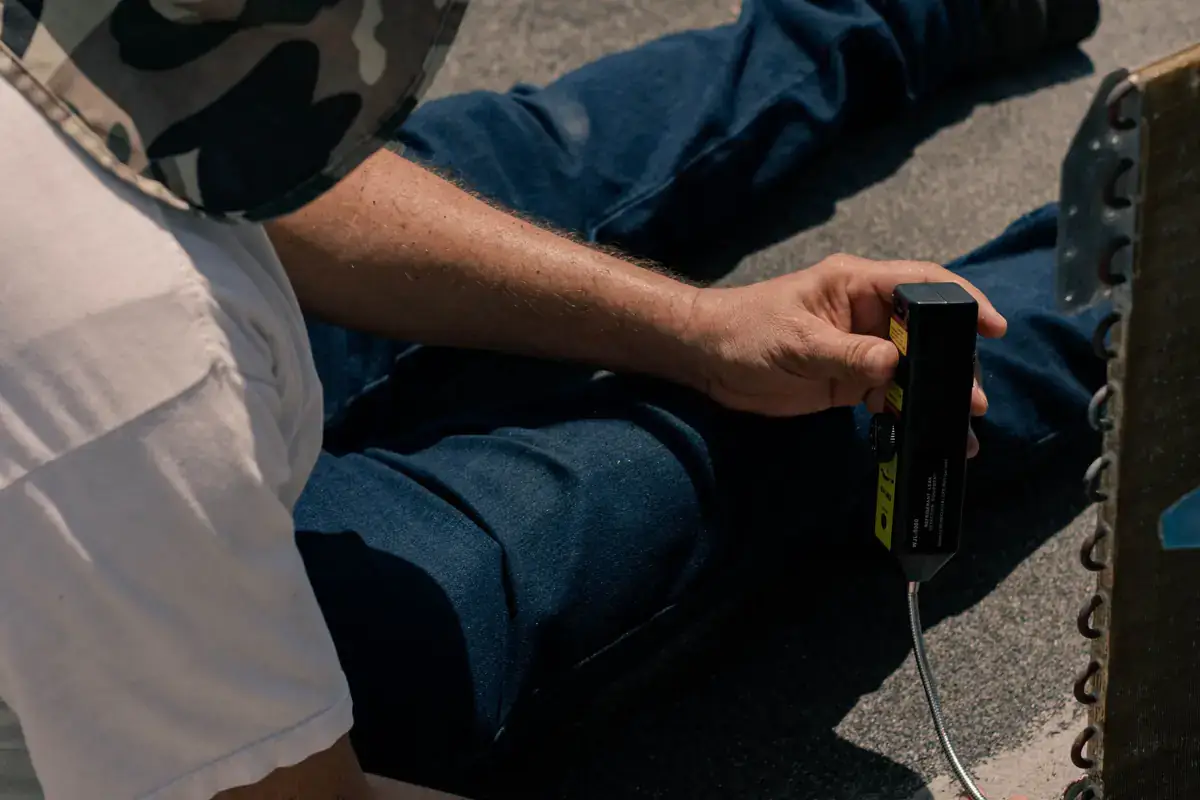 Technician seated on a roof holding a handheld tester near the coil of an air conditioning unit